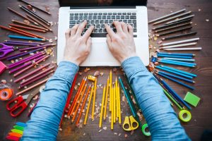 A person works on a laptop computer surrounded by coloured pencils