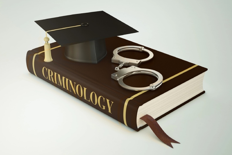 Brown book with “criminology” written post img Brown book with “criminology” written on the side which has handcuffs and a mortarboard sitting on top of it.